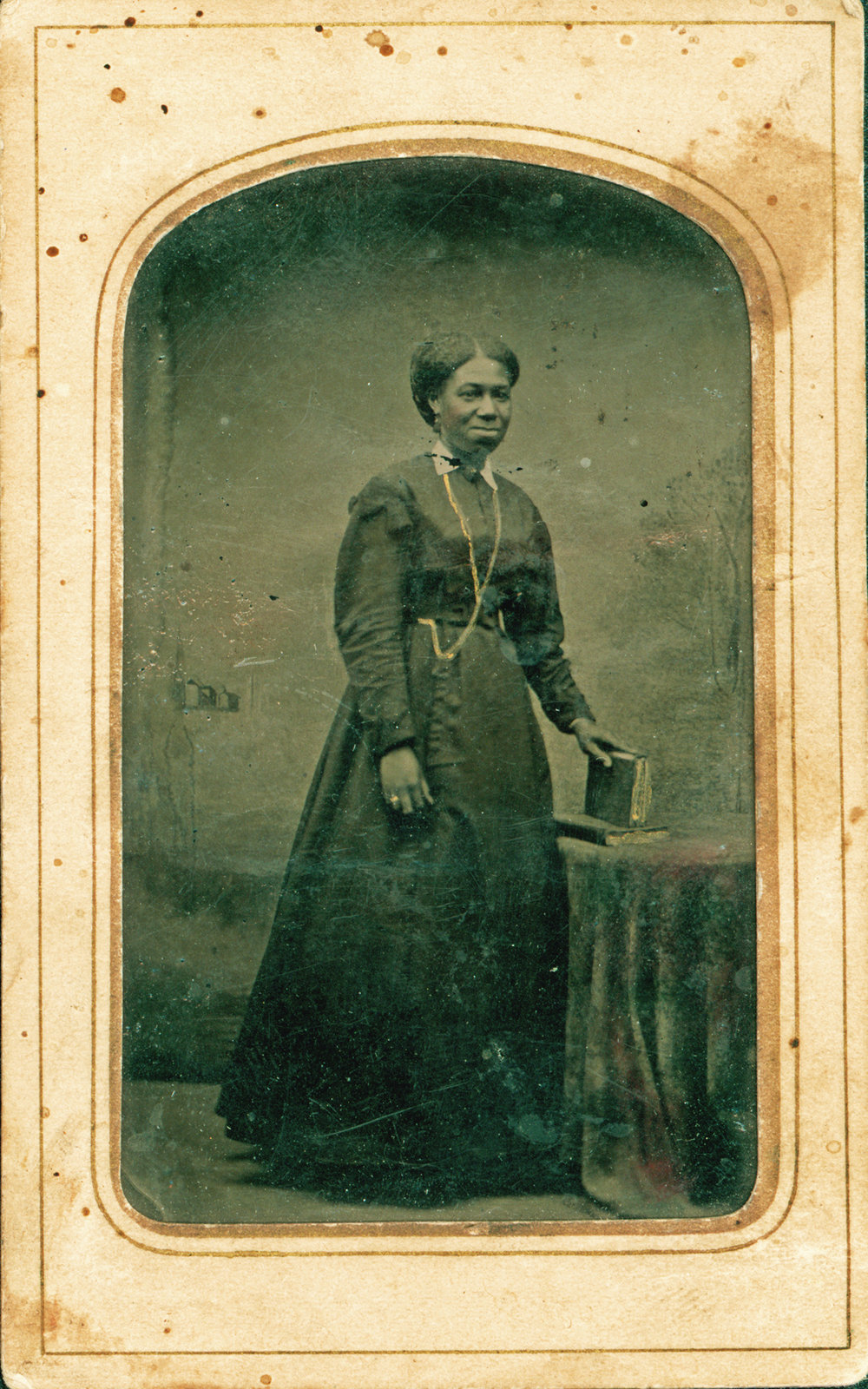 A woman standing with a book beside a table covered with a decorative cloth is featured in this small black and white tintype photograph. Courtesy the Brock University James A. Gibson Special Collections & Archives.