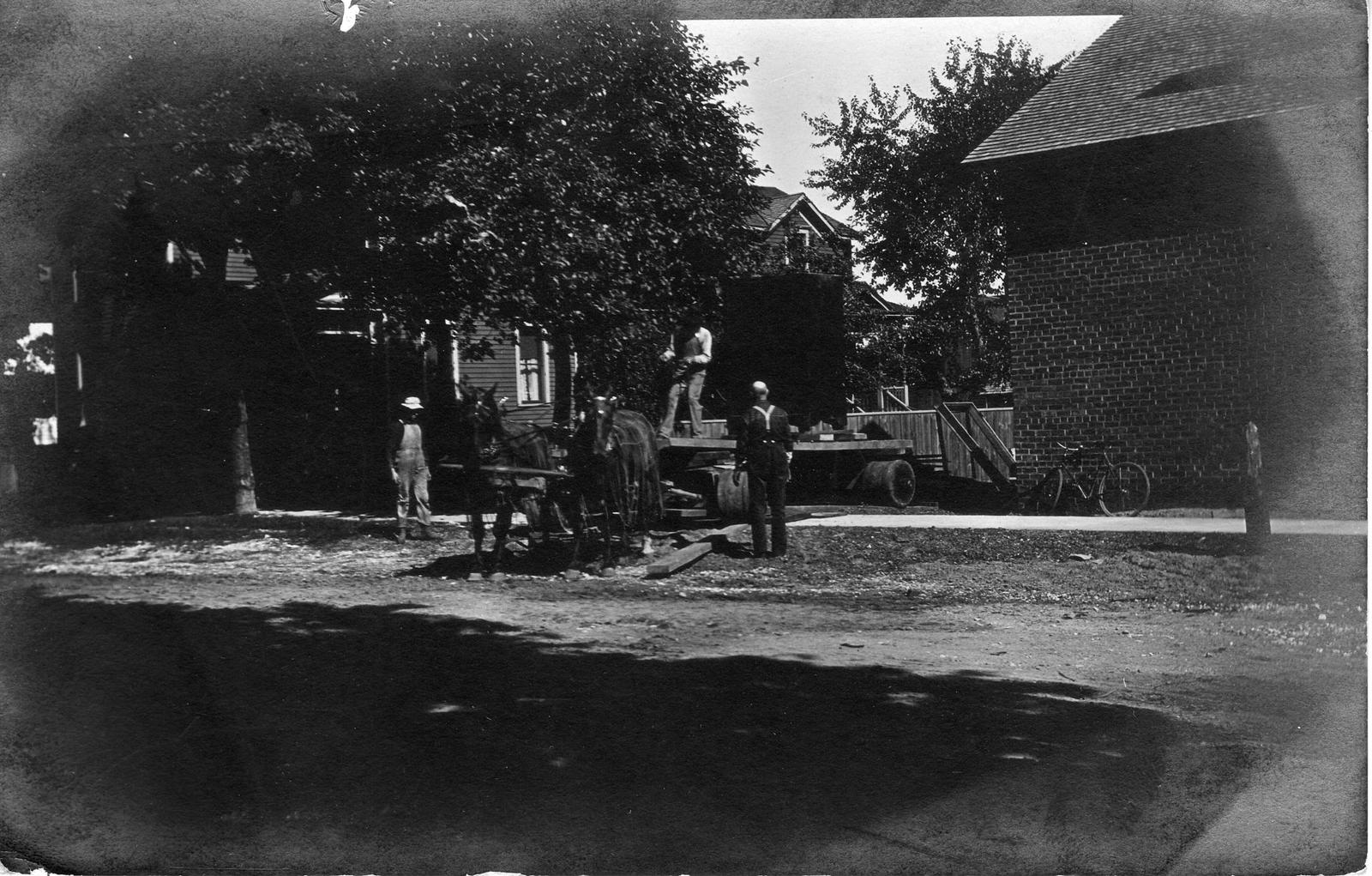 019 Three men working in a residential area with a horse team and open wagon with what looks to be a large safe. Wagon has oversized, heavy-duty wheels.