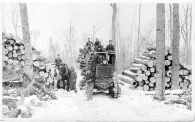 054 Crew of lumbermen with snow tractor and team of horses and large piles of logs in woods