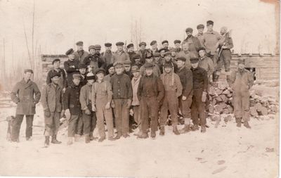 064 Crew of 40 men standing outside lumber camp building in winter