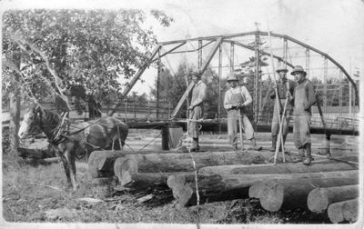 235 Four  Native American men standing on logs pulled from a river by a horse near an iron bridge.