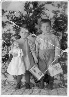 451 Two unidentified toddlers standing in front of ever green branches and holding Christmas cards and a porcelain doll. c 1900s