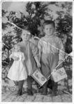 451 Two unidentified toddlers standing in front of ever green branches and holding Christmas cards and a porcelain doll. c 1900s