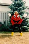 Applefest - Apple mascot 1984 standing in front of fence and a blue spruce.