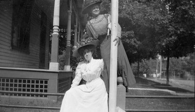 Two woman posing on a fence by a flagpole in front of a house