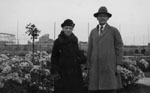 Man and elderly lady standing in front of flowering shrubs.