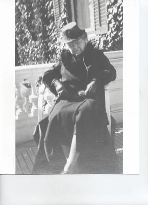 Older woman sitting in a chair on a patio outside. She is wearing black or dark coloured garments and a hat.