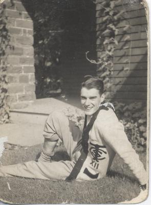 A young man relaxing on a lawn, wearing what appears to be a cardigan from a school uniform. The cardigan displays the letters &quot;T C S&quot;.