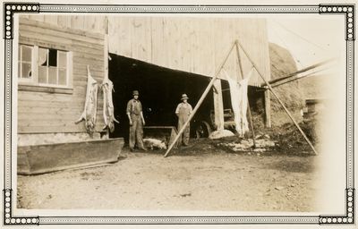 Two men standing beside a house, under an overhang with pig/animal carcasses