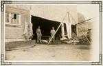 Two men standing beside a house, under an overhang with pig/animal carcasses