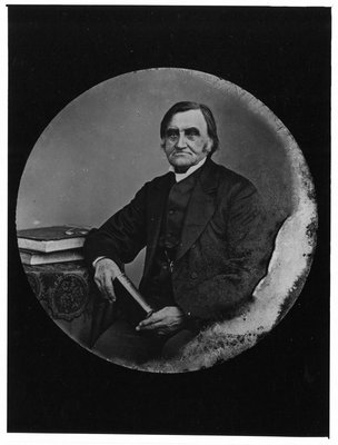 Portrait of an old man holding a book, seated at a table.