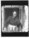 Portrait of an elderly woman standing in a doorway, behind a picket fence.