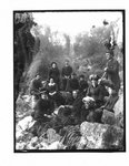 Group portrait of unidentified men and women on a rocky river bank with a bridge over the Irvine river (Elora) in the background.