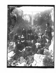 Group portrait of unidentified men and women on a rocky river bank with a bridge over the Irvine river (Elora) in the background.