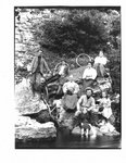 Portrait of a group of men, women and children, some with bicycles, seated on a rocky river bank.