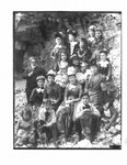 Portrait of a group of women and children, along with one man, and a dog, on a rocky river bank.