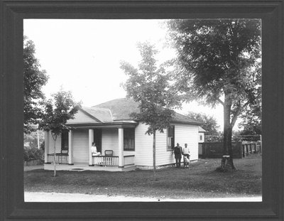 A white clapboard cottage where a woman sits reading on the front porch and a man, boy and dog pose at the side of the house.