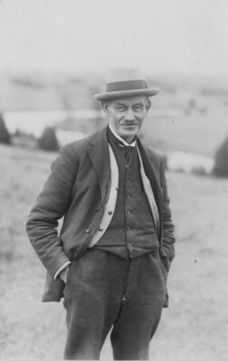 Outdoor portrait of an unidentified man,wearing felt hat, dark waistcoat, light coloured cardigan and dark jacket and trousers.