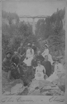 Group of unidentified men and women posed on a rocky bank, with the bridge over the Irvine River, Elora, in the background.