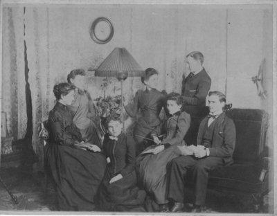 Group portrait of 2 men and 5 women in a living room/parlour.