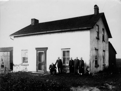 Group portrait of 5 elderly people [3 men, 2 women], in front of a storey and half stucco house.